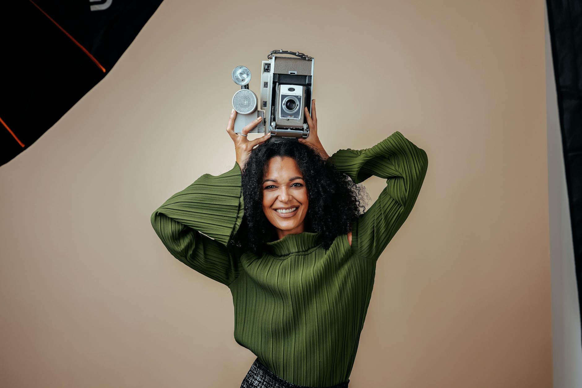 Cheerful woman in green holds a retro camera in an indoor studio setting.