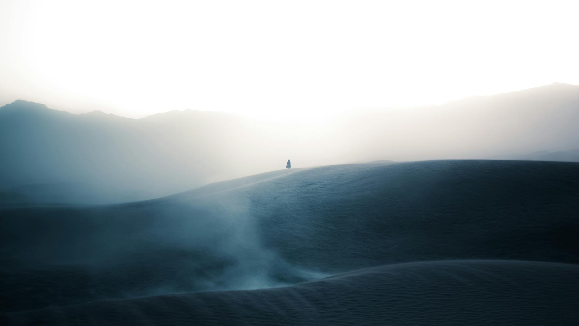 A lone figure stands silhouetted in misty desert dunes at sunrise, creating a serene and mysterious atmosphere.