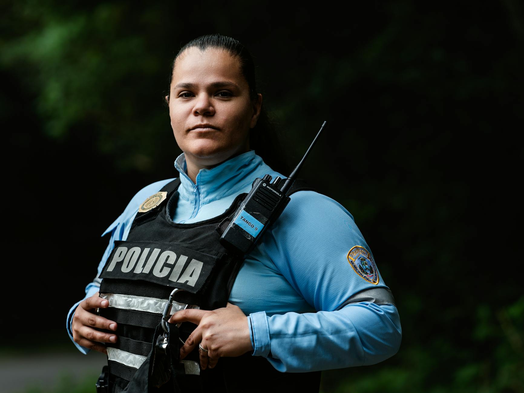 Female police officer in uniform standing outdoors with a confident expression and radio handset.