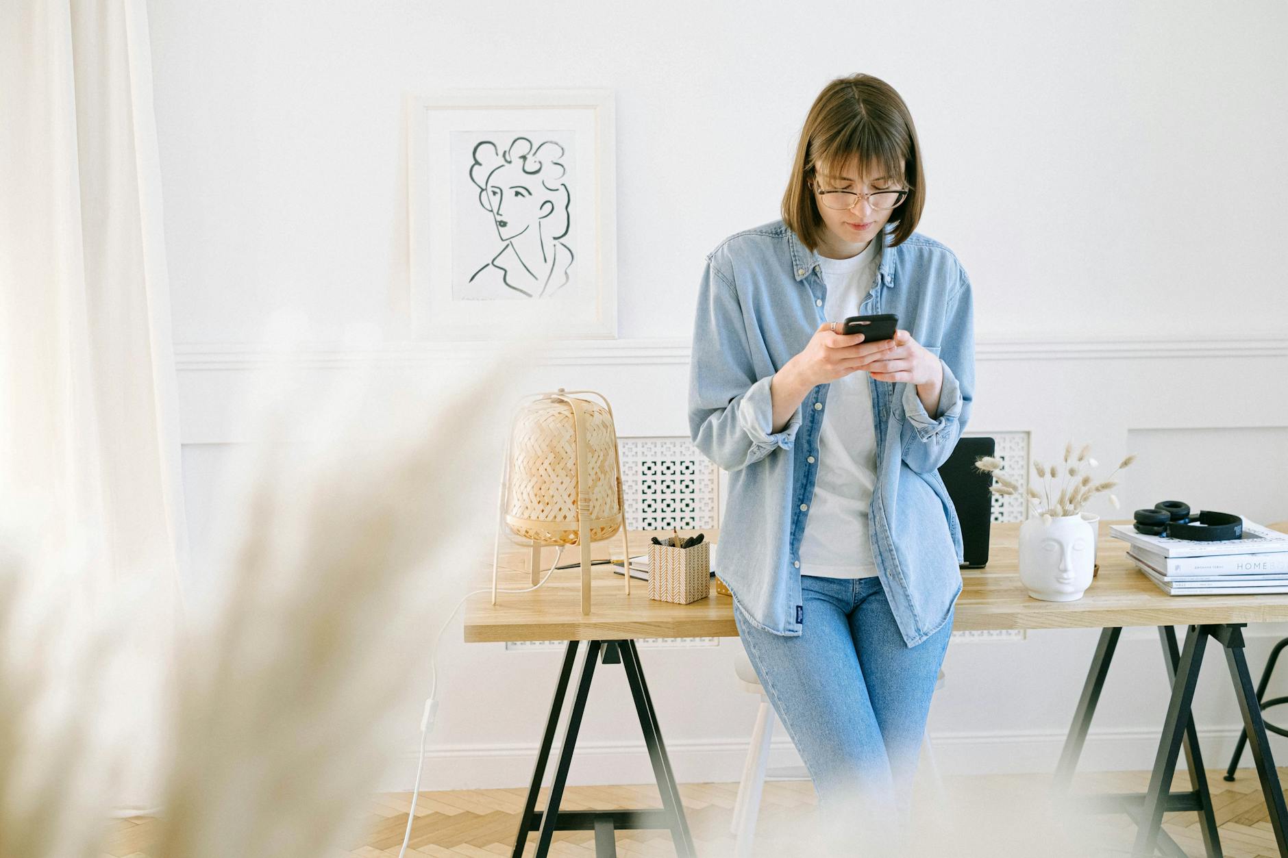 Woman in denim using smartphone in a bright home office setting.