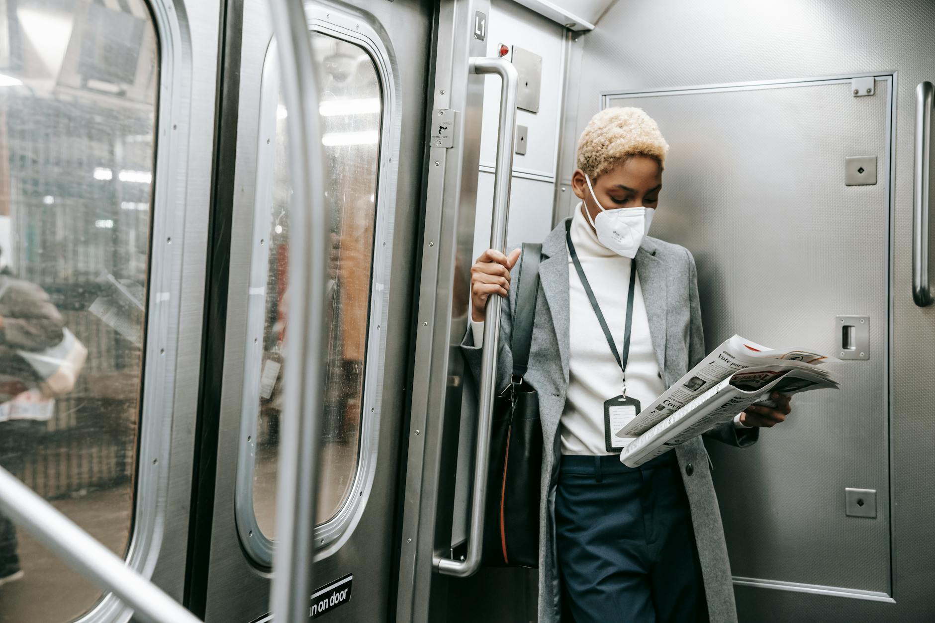 Concentrated young African American female in formal outfit and protective mask reading fresh newspaper while standing in metro train corner