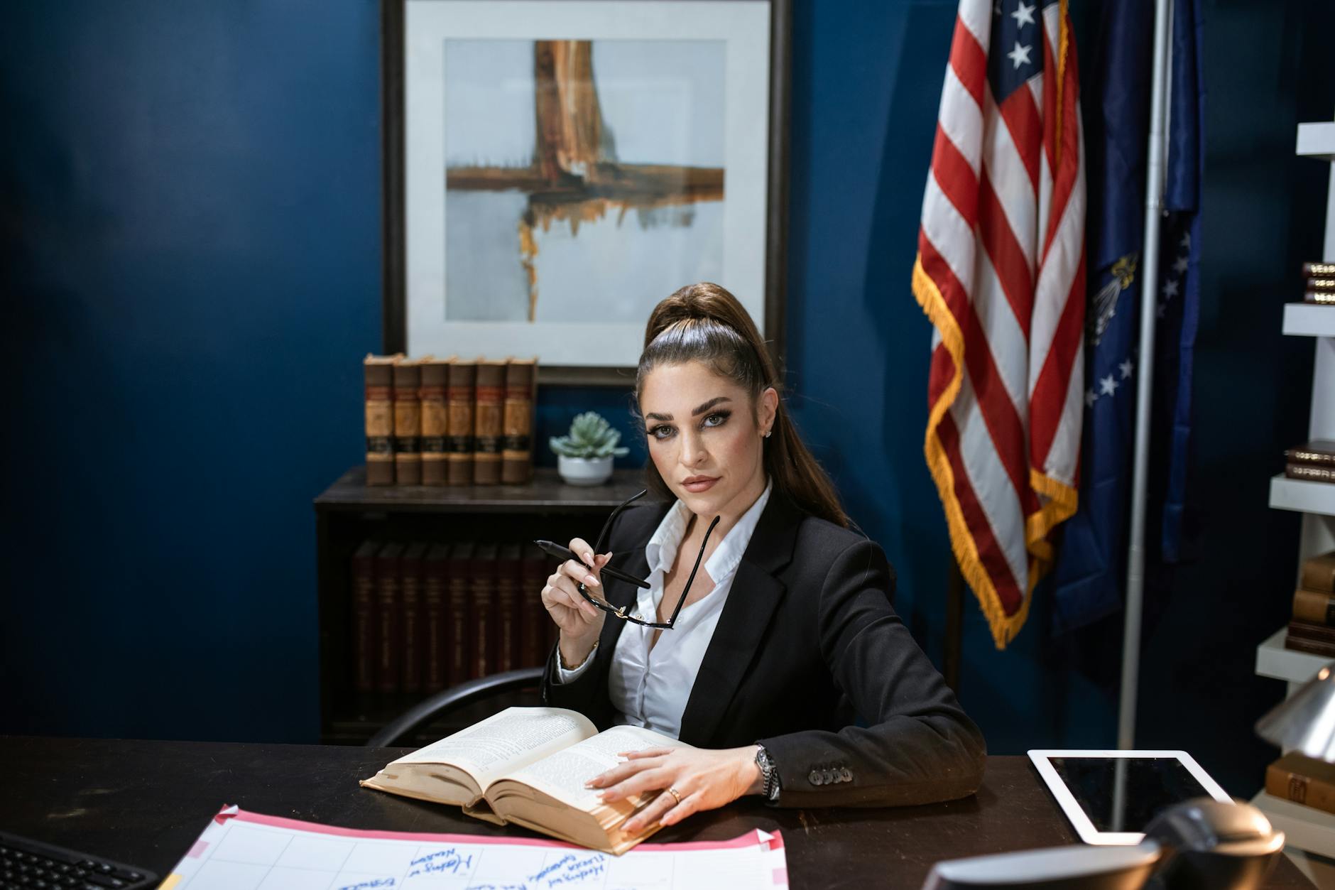 Businesswoman in black blazer and white shirt holds eyeglasses, sitting at a desk with books.
