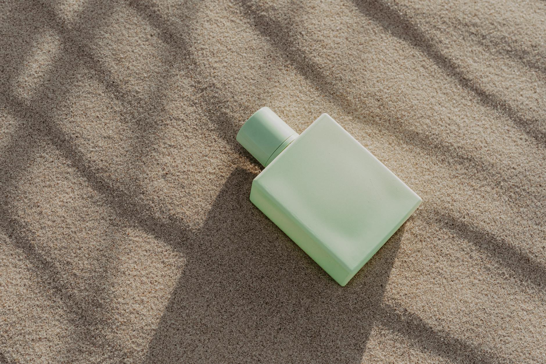 A minimalist perfume bottle lies elegantly on sand, casting soft shadows in natural light.