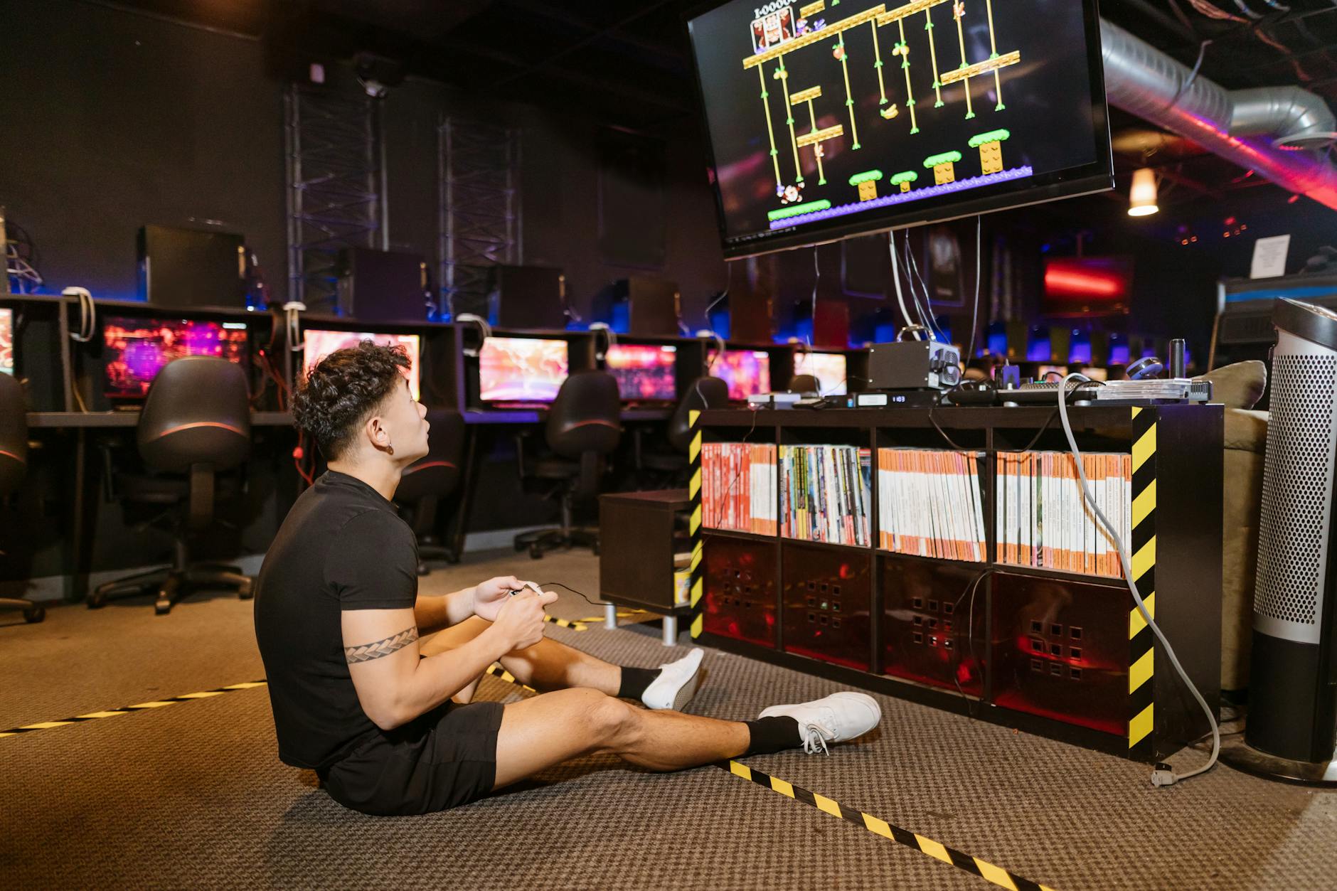 Man sitting on floor intensely playing video game at a modern arcade with vibrant computing equipment.
