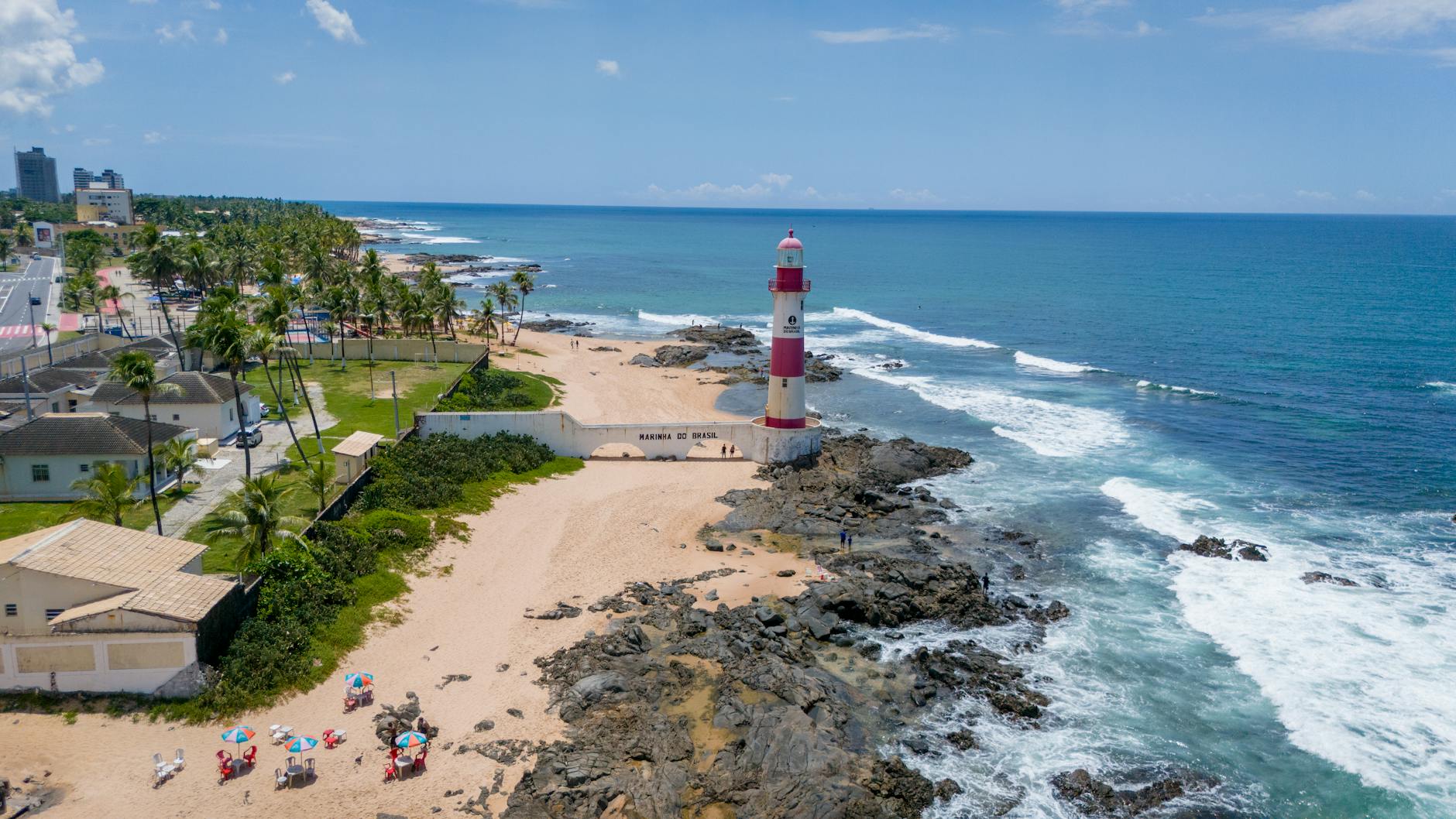 Aerial shot of Itapuã Lighthouse on a sunny beach in Salvador, Brazil, with waves crashing on the rocks.
