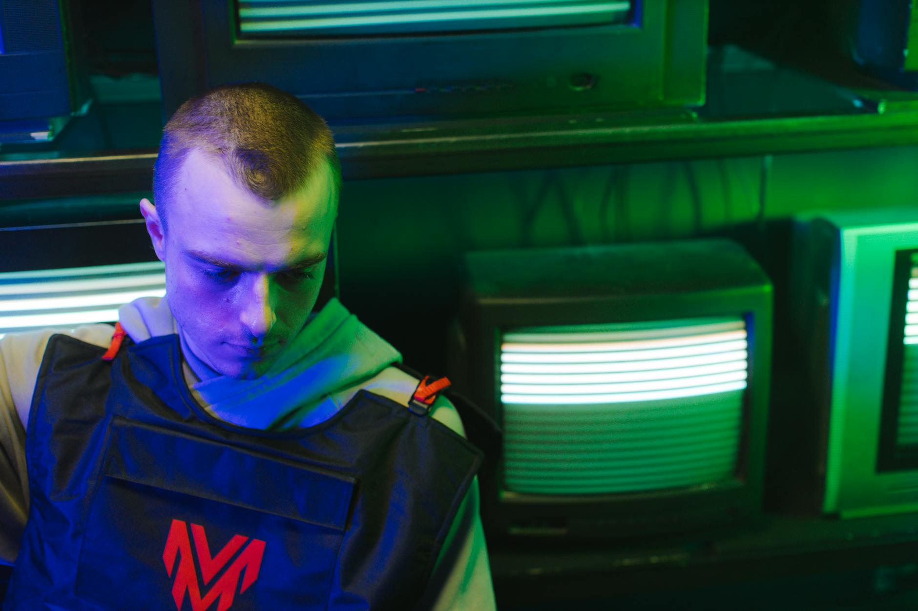 A man dressed in a vest and hoodie is surrounded by illuminated vintage CRT monitors in a cyberpunk setting.