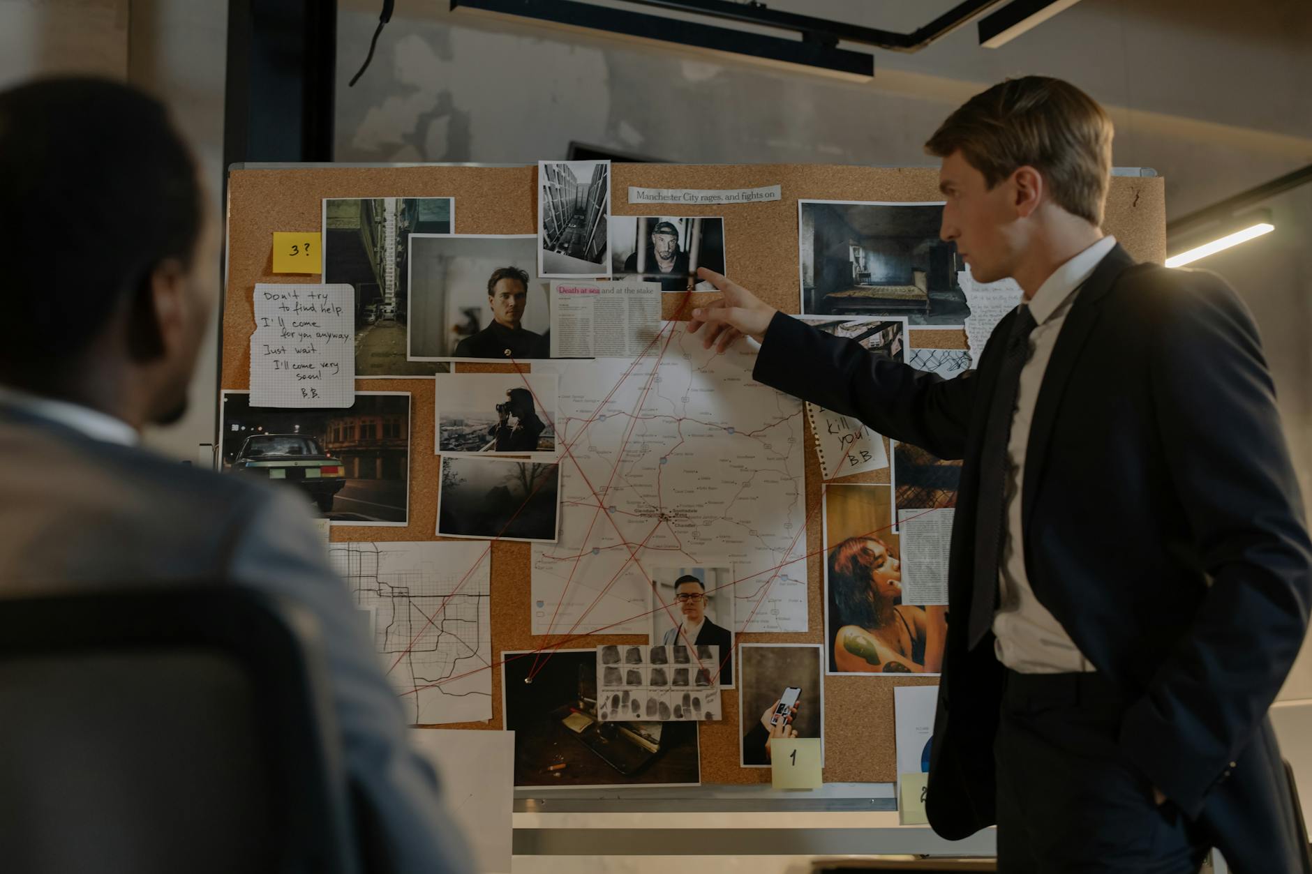 Two detectives discussing suspects and evidence on a cork board in an office setting.